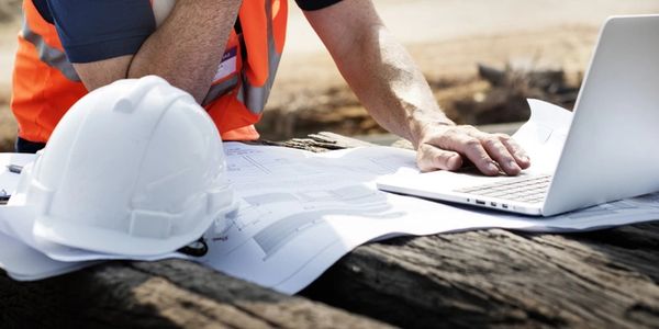 Construction worker reviewing plans with laptop and helmet on site.