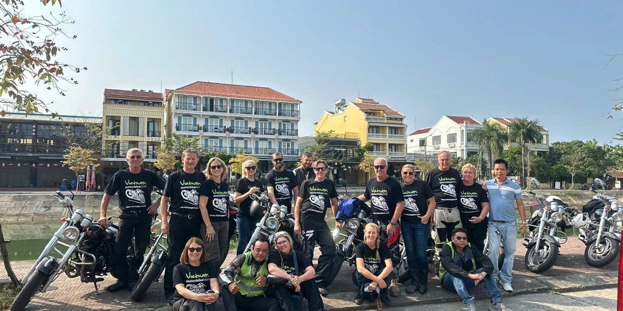 Group of motorbike riders posing together in front of motorcycles on a sunny day.