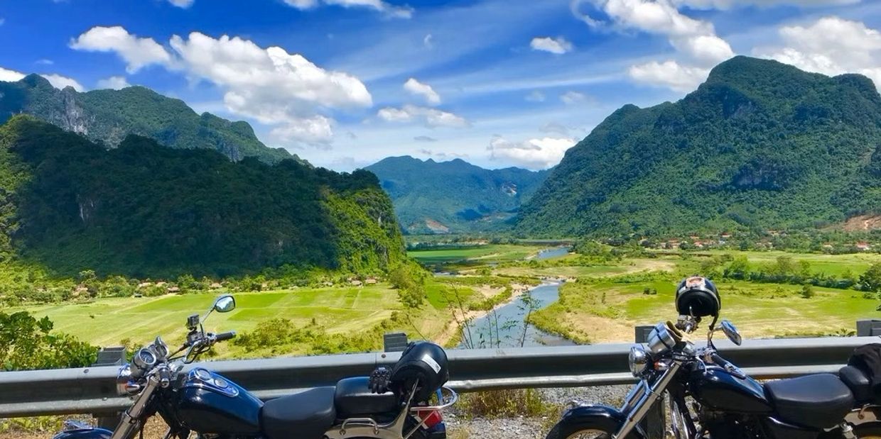Two motorcycles parked with a scenic mountain and river backdrop under a blue sky.