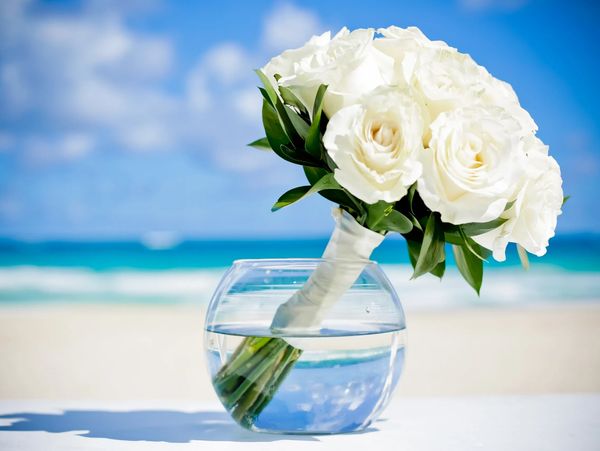 A bouquet of white roses in a clear round vase on a beach table.