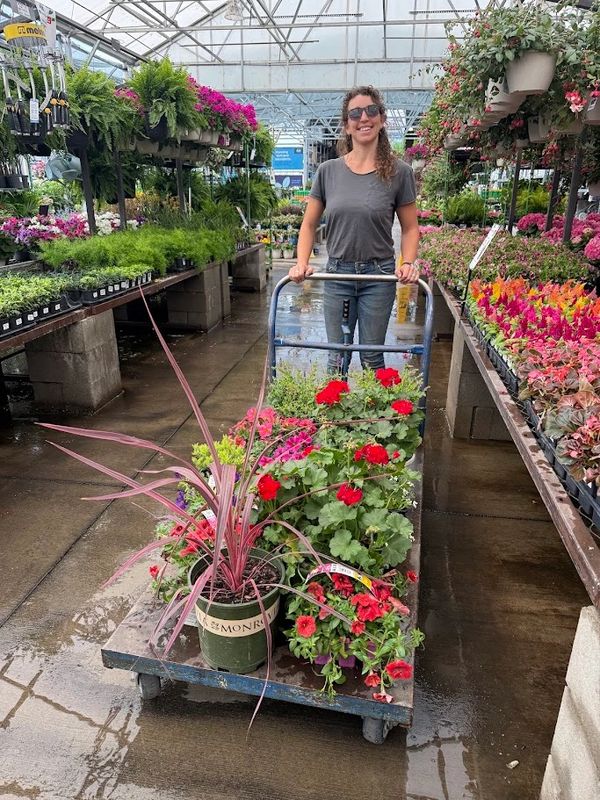 Woman smiling with cart full of colorful plants in a greenhouse.