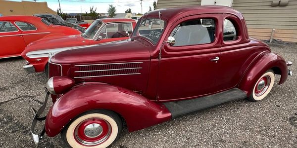 A vintage maroon classic car parked on a gravel lot with other classic cars.