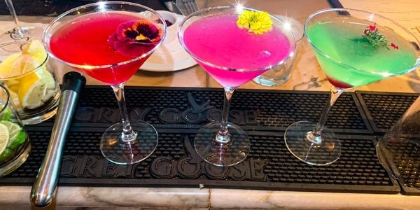 Three colorful cocktails with flower garnishes at a bar.