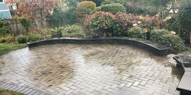 A wide, curved patio made of interlocking grey pavers, bordered by a low stone wall and garden lands