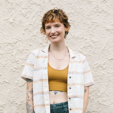Kendall, a person with short orange-ish hair, tattoos and a crop top & standing against a wall. 