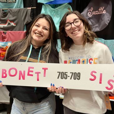 Two smiling women holding a sign for Bonett Sisters booth.