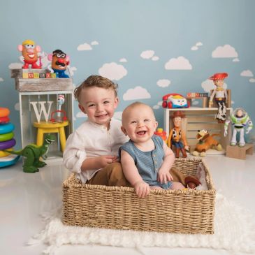 Two smiling children sitting in a wicker basket surrounded by colorful toys.