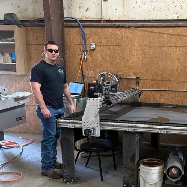 Man wearing sunglasses stands next to industrial machinery in a workshop.