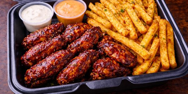 Barbecue glazed chicken wings with seasoned fries and two dipping sauces in a takeout container.