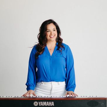 Woman in blue blouse standing behind a Yamaha keyboard, smiling.