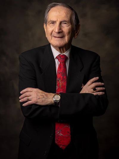 Elderly man in a suit with red tie, arms crossed, smiling.