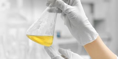 Tubo de ensaio cônico com líquido amarelo segurado por mãos com luvas brancas em um laboratório.