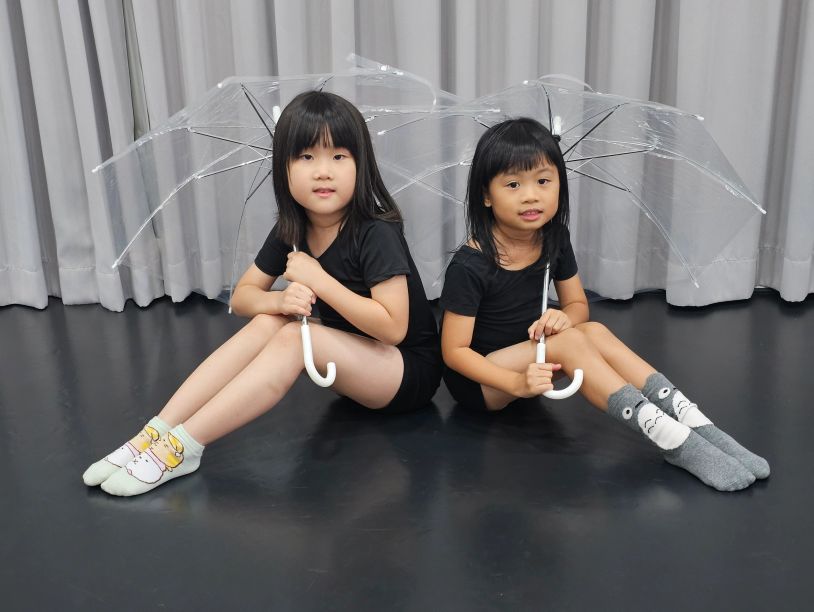 Two girls sitting back-to-back holding clear umbrellas indoors.