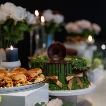 A green cake with chocolate drip and mini sandwiches on a table with candles and flowers.
