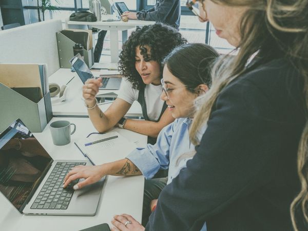 Three women collaborating on a laptop in a modern office space.