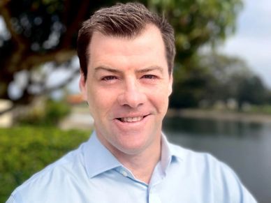 Smiling man in a light blue shirt outdoors near water and greenery.