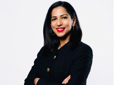 Confident woman with black hair and red lipstick, wearing a black textured jacket with gold buttons, smiling with arms crossed.
