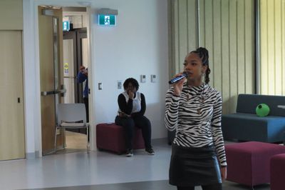 Woman speaking into a microphone in a modern indoor space with people sitting in the background.