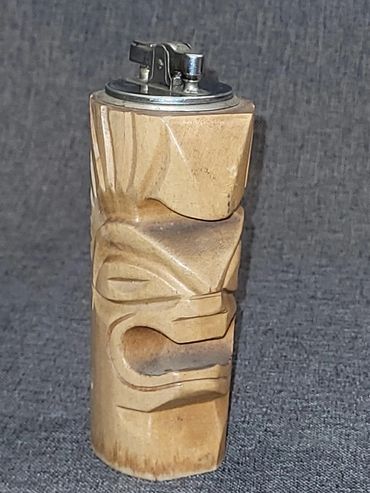 Carved wooden tiki lighter with a silver top against a gray background.