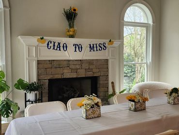 A festive banner reading 'CIAO TO MISS' hangs above a decorated fireplace in a cozy dining room.