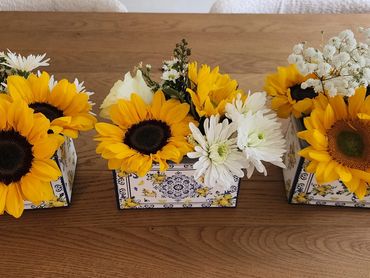 Three floral arrangements with sunflowers and white flowers on a wooden table.