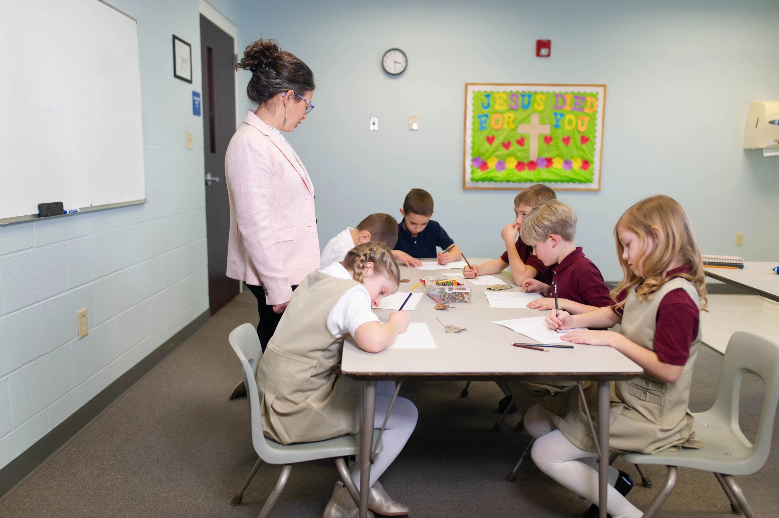 Teacher supervises children drawing at a table in a classroom with a religious poster.