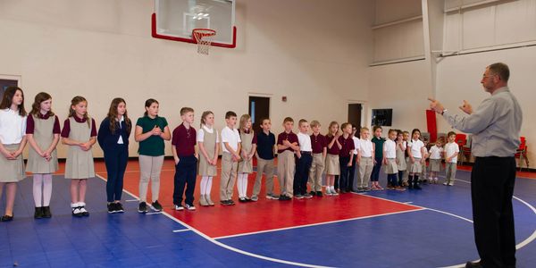 Children lined up in a gym listening to an adult giving instructions.
