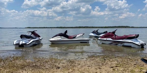 Four jet skis floating on calm lake water near a sandy shore under a cloudy sky.