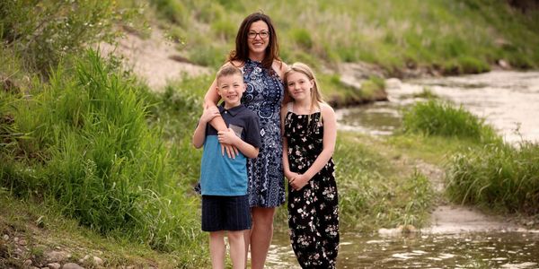 A woman stands on a river bank with two children, a boy on the left and a girl on the right.