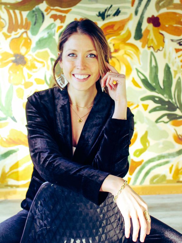 Smiling woman in black outfit sitting on a chair with floral background.