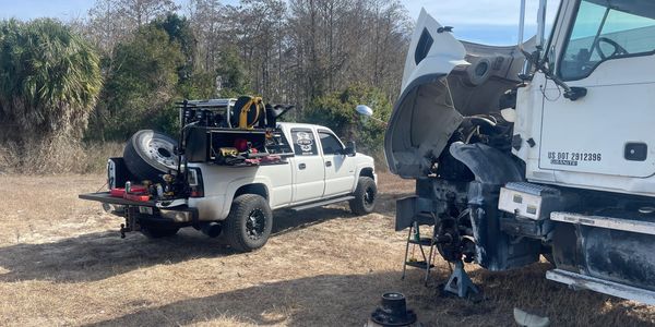 A white pickup truck parked near a large truck undergoing repairs in a grassy area.
