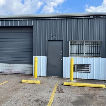 Gray industrial building with a garage door, entrance door, barred window, and yellow parking barriers.