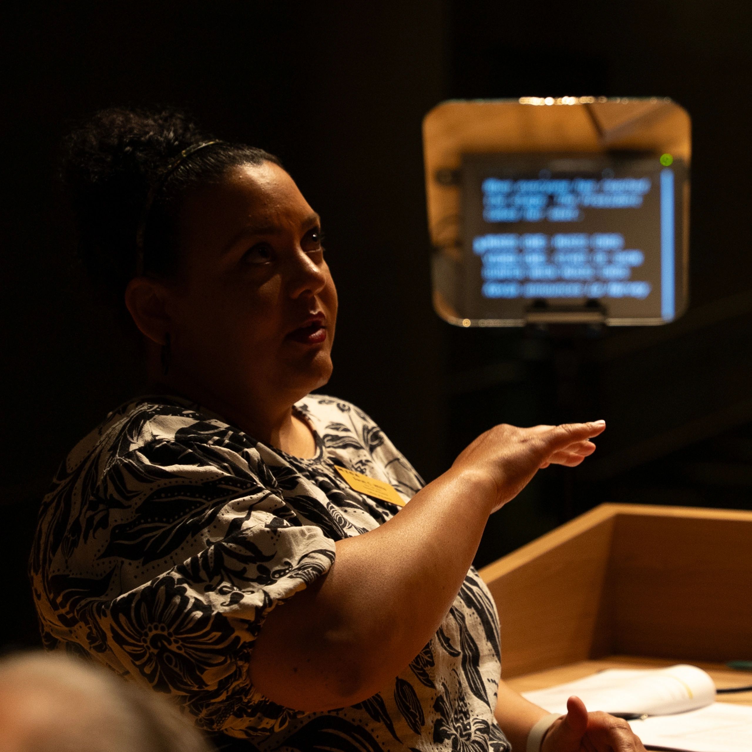 Woman speaking at a podium with a presidential teleprompter in the background.