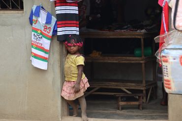 A young child stands barefoot at a doorway, surrounded by hanging clothes.