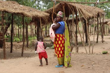 A woman carrying a baby with a child nearby in a rural setting.