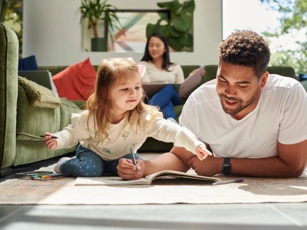A father and daughter draw together on the floor while a woman works in the background.