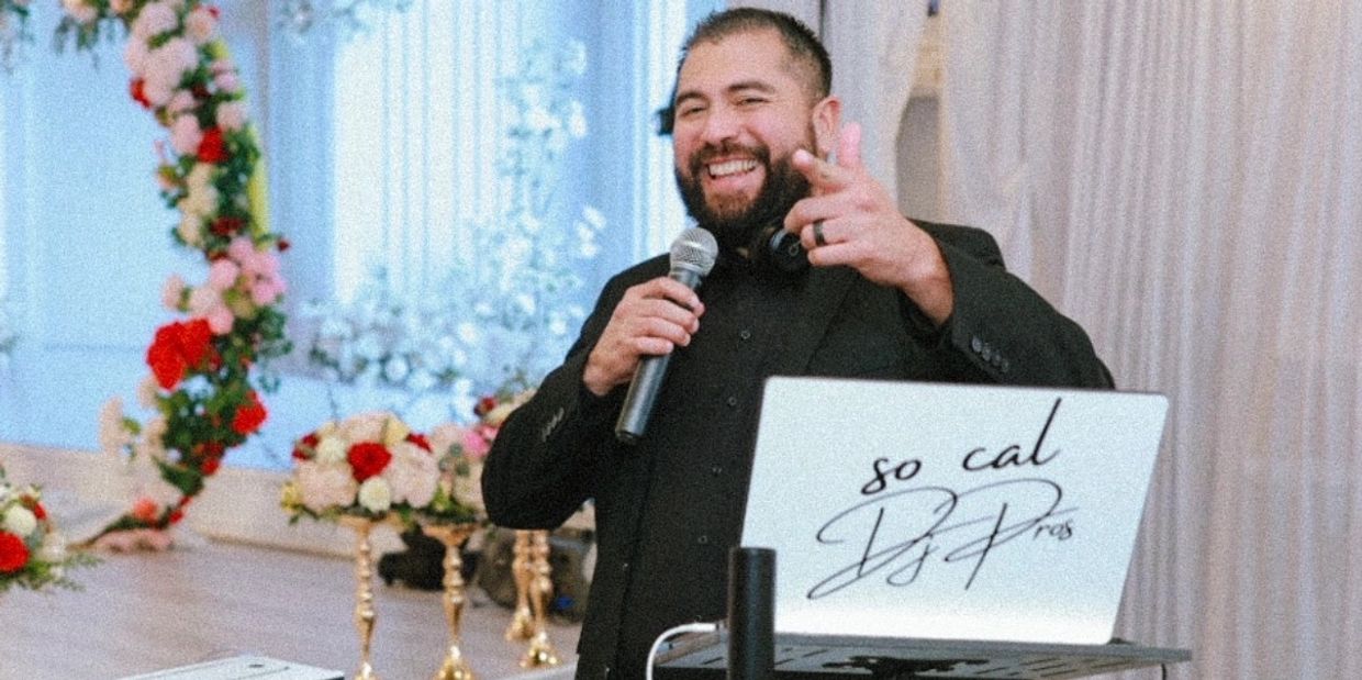 A joyful DJ holding a microphone and pointing, standing behind a laptop with floral decorations in the background.