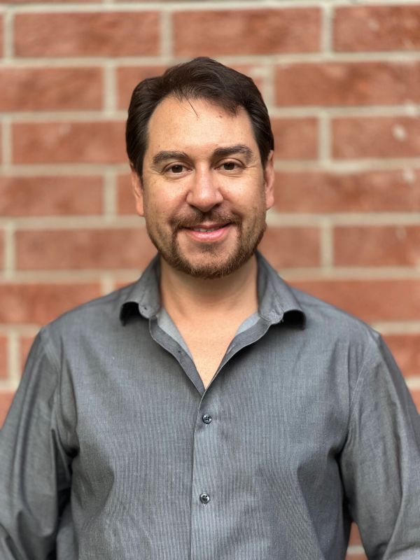 Smiling man in gray shirt against a brick wall.
