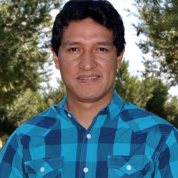 Man in a blue plaid shirt posing outdoors with trees in the background.