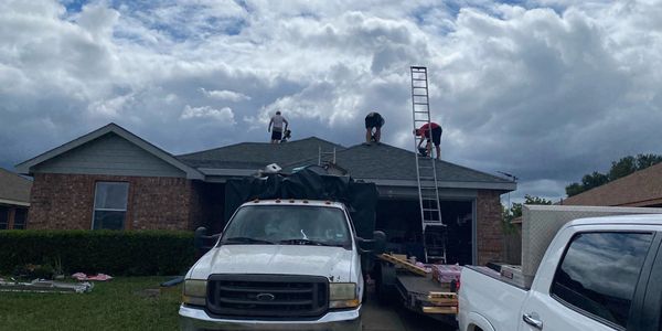 Workers repairing the roof of a brick house with trucks parked in the driveway.