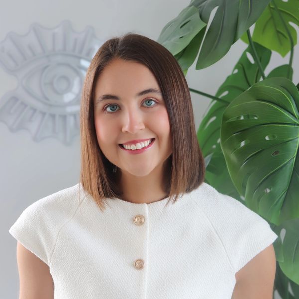 Young woman smiling with large green leaves and eye decor in background.