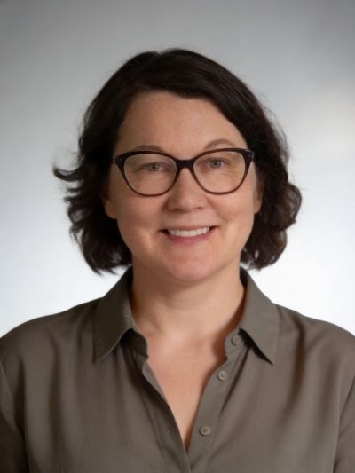 Woman with dark hair and glasses smiling against a plain background.