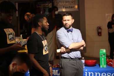 Two men engaged in conversation at an indoor event with a table of items nearby.