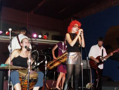 Band performing live with a singer in a red wig and musicians playing various instruments.