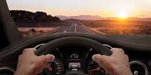 View of a driver holding a steering wheel with a sunset ahead.