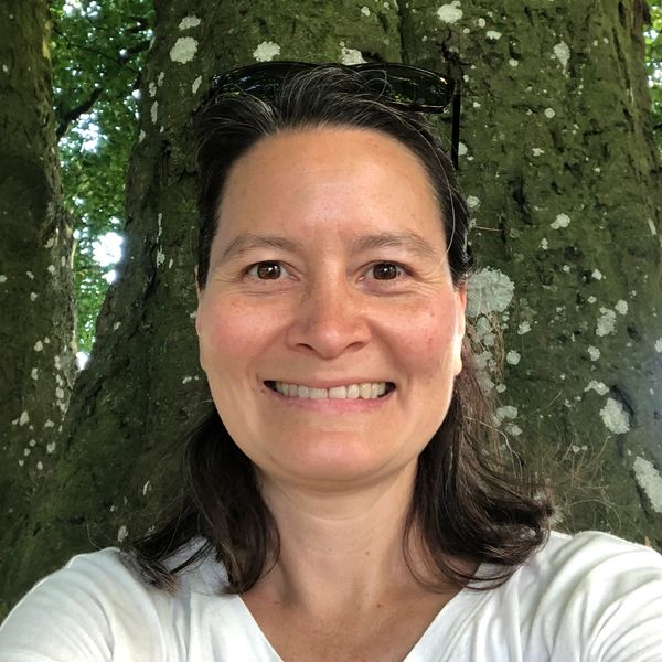 Smiling woman taking a selfie outdoors with a large tree in the background.