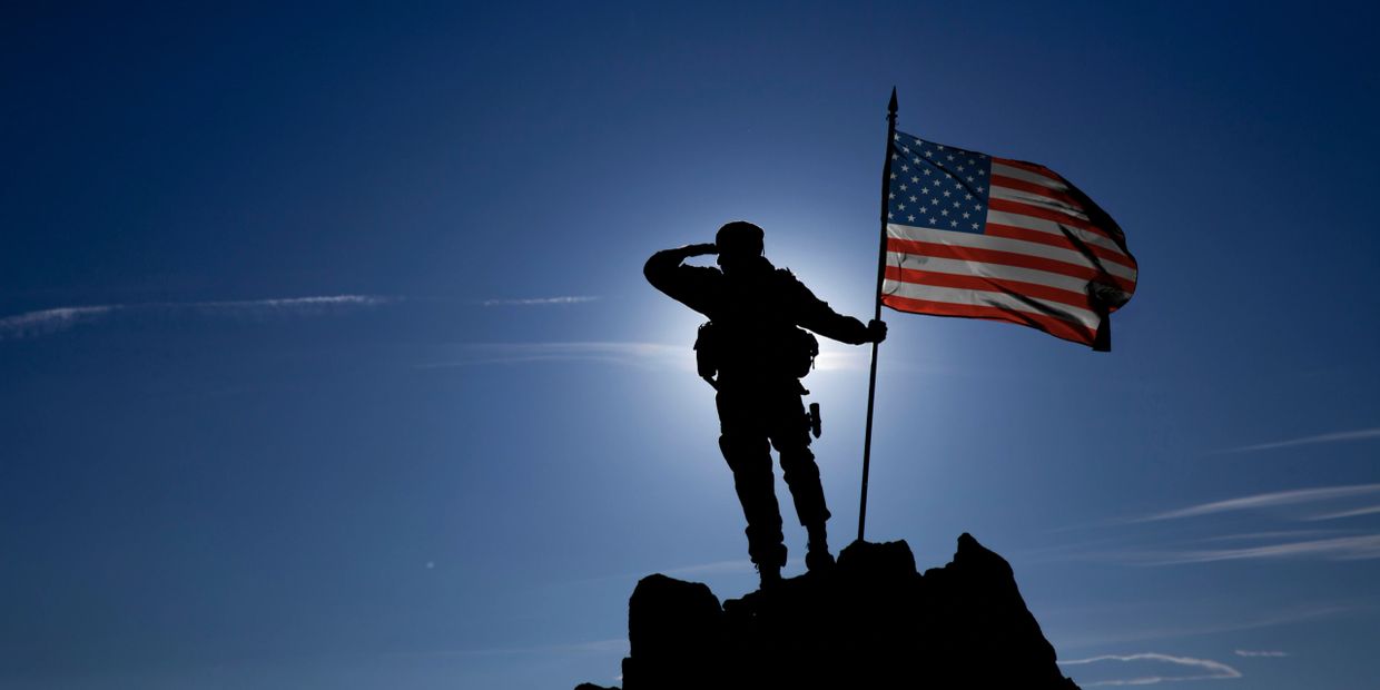 Silhouette of soldier saluting next to an American flag on a mountaintop.