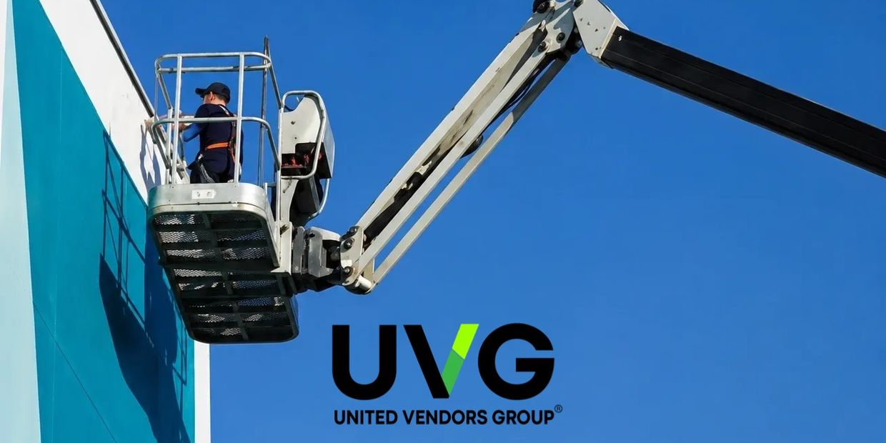 Worker on a boom lift painting a blue wall under clear sky with UVG logo.