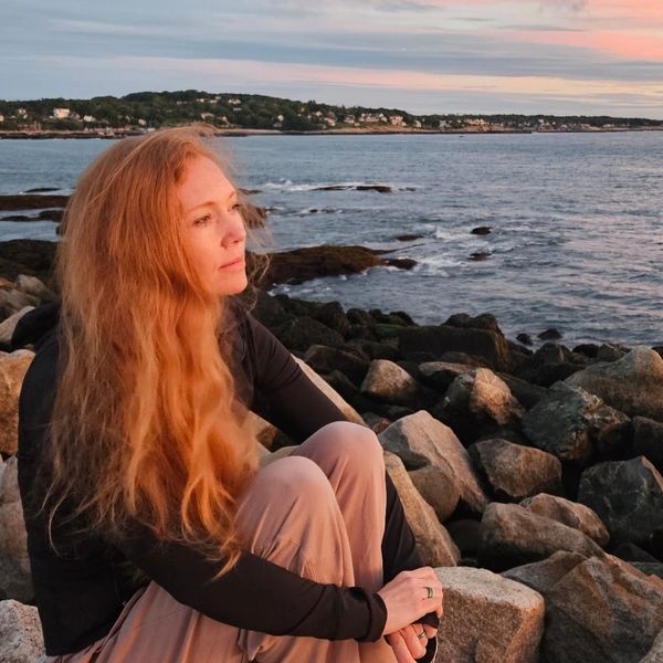 Red haired woman sits on a jetty looking out at the ocean and pink and blue sunrise.