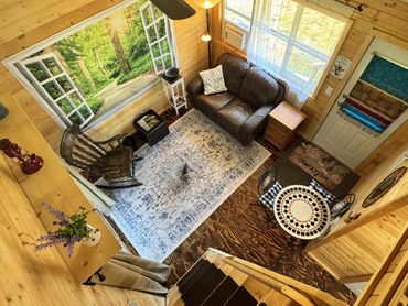Cozy cabin living room with futon, rocking chair, and large windows overlooking the forest.